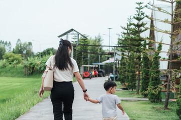 Asian mother and son walking hand in hand on a peaceful path during a family outing. The scene shows lush green nature, casual parenting, and quality bonding time in an outdoor travel setting.