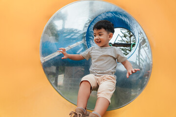 Happy Asian boy sliding down a transparent playground tunnel with a big smile. Child enjoying outdoor physical play, developing balance, confidence, and coordination through joyful movement.
