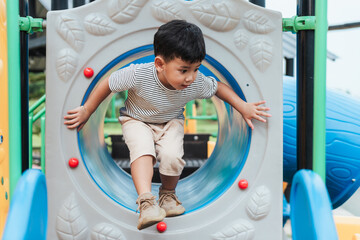 Asian boy playing and climbing out of a tunnel at outdoor playground. Developing gross motor skills, focus, self-confidence, and independence in childhood through active play and exploration.