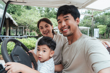 Happy selfie Asian family sitting in a golf cart with toddler holding the steering wheel. Parents smiling during zoo trip. Real candid moment of travel, parenting, bonding, and fun family experience.