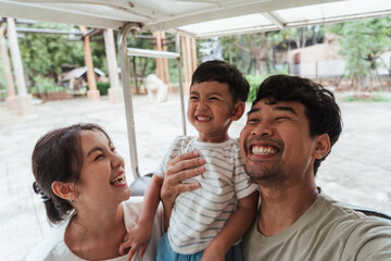 Smiling Asian family taking a selfie on golf cart during zoo visit. Parents holding joyful toddler with big laugh. Real travel lifestyle moment, full of love, bonding, and authentic family happiness.