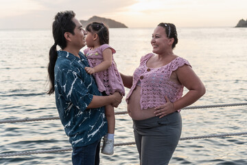 Hispanic family enjoying sunset at the beach, pregnant mother and father holding daughter
