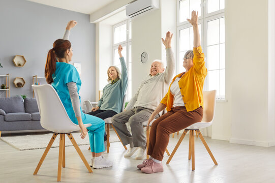 Young nurse leading seated exercise session with elderly patients in nursing home. Group of happy senior people sitting on chairs, raising their arms during physical therapy for mobility and wellness.