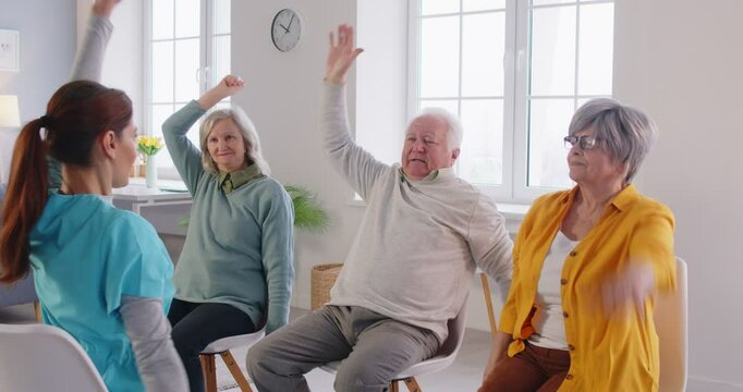 Group of seniors sitting on chairs in retirement home and doing sport exercises for health with young nurse. Elderly people working out in fitness class of nursing home. 4k video. Slow motion video.