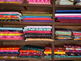 Colorful floral and solid fabric stacks neatly organized on old wooden shelves exhibit a diverse...