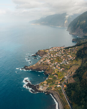Aerial view of rugged cliffs meet the deep blue sea, cradling a quaint village under a soft, hazy sky, Seixal, Setubal, Portugal.