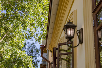 A decorative outdoor wall lantern is mounted on a building facade surrounded by green foliage,...