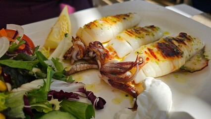 Grilled full squids calamari on the white plate with lemon and green salad leaves on the side 