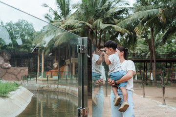Asian mother carrying her young son as they observe animals behind glass at the zoo. Reflections on...
