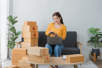 E-commerce Hustle: Young Asian entrepreneur managing orders and logistics on phone and tablet, surrounded by shipping boxes in her home office.	