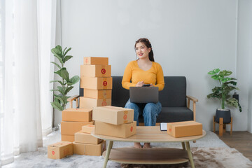E-commerce Hustle: Young Asian entrepreneur managing orders and logistics on phone and tablet, surrounded by shipping boxes in her home office.	