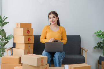 E-commerce Hustle: Young Asian entrepreneur managing orders and logistics on phone and tablet, surrounded by shipping boxes in her home office.	