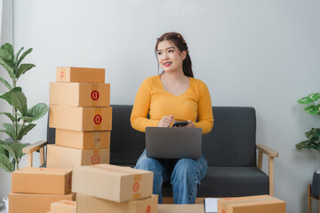 E-commerce Hustle: Young Asian entrepreneur managing orders and logistics on phone and tablet, surrounded by shipping boxes in her home office.	