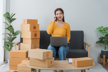 E-commerce Hustle: Young Asian entrepreneur managing orders and logistics on phone and tablet, surrounded by shipping boxes in her home office.	