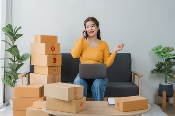 E-commerce Hustle: Young Asian entrepreneur managing orders and logistics on phone and tablet, surrounded by shipping boxes in her home office.	