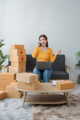 E-commerce Hustle: Young Asian entrepreneur managing orders and logistics on phone and tablet, surrounded by shipping boxes in her home office.	
