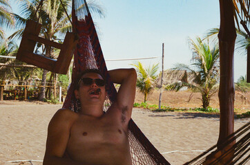 A young man enjoys a cigar in sunny weather while lounging in a hammock, with a tropical beach backdrop including palm trees and sandy terrain, representing leisure, vacation, and tranquil outdoors.