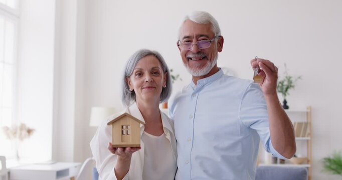 Happy senior smiling couple moving new flat, holding toy house showing keys from new own house, getting bank loan for older citizen, elder family making deal with property real estate purchase dream