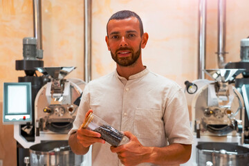 A skilled worker holds a container of freshly roasted arabica and robusta coffee beans in a roasting workshop, surrounded by advanced roasting machines, focusing on the art of coffee preparation