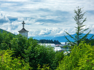 A cross atop the steeple of a hillside church in the port town of Wrangell, Alaska, with a view of distant mountains, some capped with snow, on a cloudy morning early in summer
