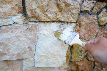 A brush applies a clear coating to an exterior natural stone wall. The wall, composed of irregularly shaped blocks, gains a deep wet sheen, enhancing the texture and color of the surface.