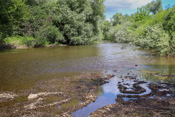A harmonious landscape displays a calm river, surrounded by dense greenery of trees and bushes. In...