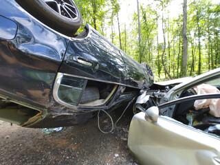 Two damaged cars involved in collision, with one vehicle flipped on its side surrounded by trees.