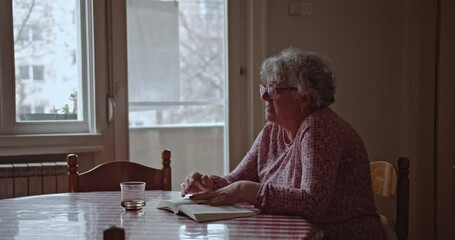 A senior woman sits at her dining table, reading a book by the window. She wears glasses and a pink patterned shirt and occasionally looks out the window. - Powered by Adobe