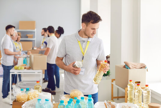 Portrait of young man volunteer sorting donations of foodstuffs in cardboard boxes for charity food drive. People volunteering in food bank working in charitable foundation together.