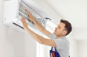 Professional electrician man in uniform maintaining or cleaning modern air conditioner indoors....