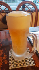 A tall, frosted glass mug filled with cold beer and a thick foamy head sits on a placemat at a...