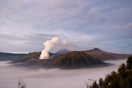 View of volcanic peaks piercing through a sea of ethereal mist, crowned by a plume of smoke against a backdrop of serene sky, Mount Bromo, East Java, Indonesia.