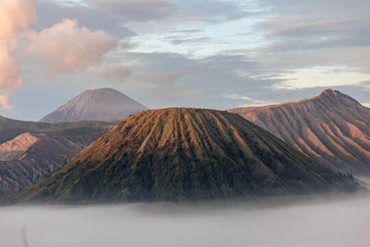 View of majestic, sunlit volcanoes piercing through a sea of ethereal clouds, creating a stunning contrast of earth and sky, Mount Bromo Volcano, East Java, Indonesia.