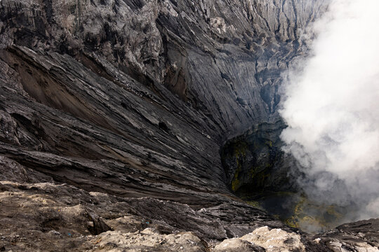 View of the volcanic crater's jagged, grey rock formations meet billowing white smoke rising from the depths, a stark contrast, Mount Bromo, East Java, Indonesia.