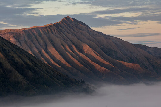 View of majestic, sun-kissed mountain ridges cascading into a sea of ethereal mist under a pastel sky, Mount Bromo Volcano, East Java, Indonesia.