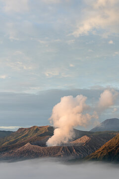 View of volcanic peaks pierce through a sea of ethereal mist, plumes of smoke rising against a backdrop of soft, pastel skies, Mount Bromo, East Java, Indonesia.