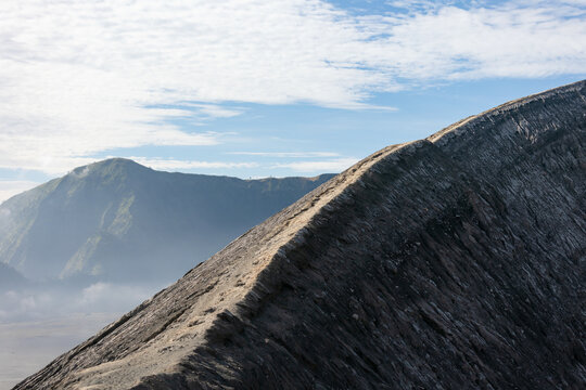 View of rugged, grey volcanic terrain rising dramatically against a backdrop of misty mountains and a bright, blue sky, Mount Bromo Volcano, East Java, Indonesia.