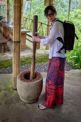 A smiling woman in a white shirt and patterned skirt uses a large traditional mortar and pestle to...
