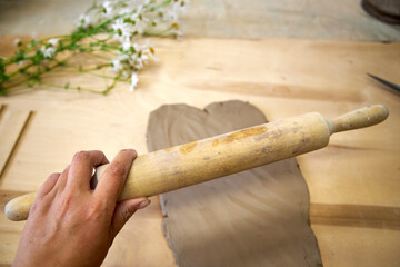 Close-up of a hand rolling out clay on a wooden surface with a rolling pin. Chamomile flowers in...
