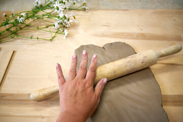 Close-up of a hand rolling out clay on a wooden surface with a rolling pin. Chamomile flowers in...