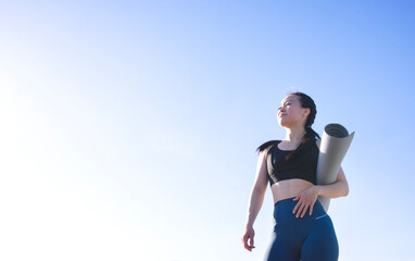 Asian woman in activewear holding yoga mat, standing outdoors against clear blue sky, embodying...