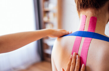 A young woman sits with her back turned, hands of a person touch her, applying kinesiology tapes....