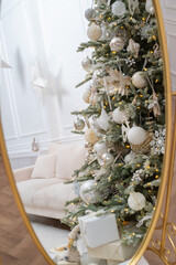 Decorated with an array of elegant ornaments in shades of white, silver, and gold Christmas tree reflected in a large, ornate mirror. Elegant interior in white and beige colors in the background.