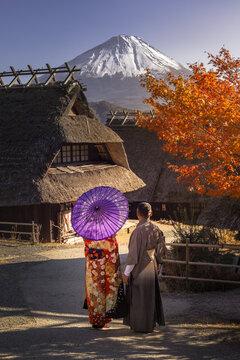 View of a couple strolling under the autumn sun, the woman with a parasol, towards traditional thatched-roof houses and snow-capped Mt. Fuji, Fuji, Shizuoka, Japan.