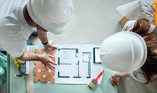 A young couple in construction helmet carefully study an architectural plan. Top view. A woman stands behind a table and look at a tablet.