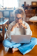 Curly-haired woman in her 40s using a laptop at home with varied expressions. Remote work, digital...