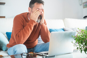 Mature man using laptop at home with mixed expressions. Remote work, digital lifestyle, emotional...