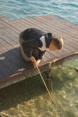 Boy bending over a wooden pier looking into the clear water, engaging in a moment of curiosity and...