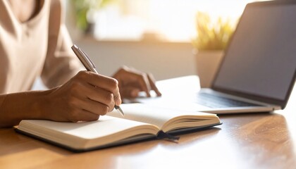 Woman's hands diligently writing in a notebook, fingers also interacting with a nearby laptop, suggesting focused work or study.