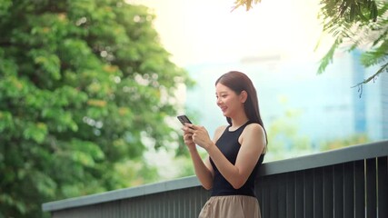 Thoughtful asian woman standing in park using smartphone while smiling gently, enjoying peaceful moment texting or browsing social media outdoors with digital lifestyle in green city environment - Powered by Adobe